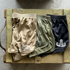 May include: Three pairs of athletic shorts in tan, green, and black with a white embroidered emblem on the side. The shorts are displayed on a wooden crate.