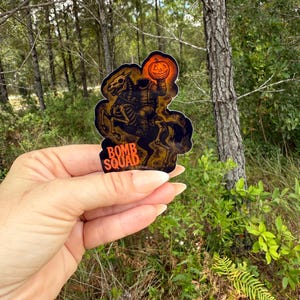 May include: A sticker featuring a headless horseman riding a skeletal horse, holding a jack-o'-lantern. The sticker has the text "BOMB SQUAD" in orange. The design is set against a dark background with orange and brown accents.