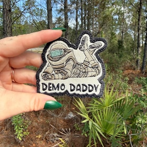 May include: Embroidered patch featuring a figure in a spacesuit-like outfit, with the text "DEMO DADDY" in white lettering. The patch has a black border and is in a forest setting.