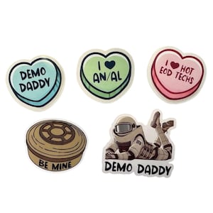 May include: Five stickers with different designs. One sticker is a heart-shaped candy with the text "I love AN/AL". Another sticker is a heart-shaped candy with the text "I love HOT EOD TECHS". A third sticker is a heart-shaped candy with the text "DEMO DADDY". A fourth sticker is a round sticker with the text "BE MINE" and a drawing of a can of food. The fifth sticker is a drawing of a person in a spacesuit with the text "DEMO DADDY".
