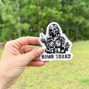 May include: Black and white sticker of a skull-faced woman in military uniform giving the middle finger. The sticker says "Bomb Squad" at the bottom.
