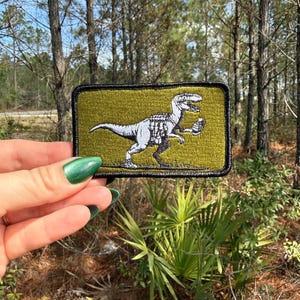 May include: A rectangular patch featuring a white dinosaur design on a green background with a black border. The patch is held in front of a forest background.