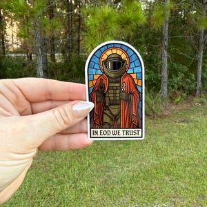 May include: A sticker depicting a stained-glass style image of a figure in a military helmet and uniform, holding a detonator. The sticker has a rectangular shape with the text "IN EOD WE TRUST" at the bottom.