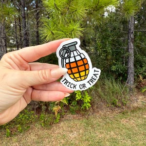 May include: A white sticker in the shape of a grenade with a black outline. The grenade is colored orange and yellow, with a black top and a silver ring. The text "TRICK OR TREAT" is written in black on a white banner.