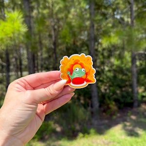 May include: A vibrant sticker depicting a cartoon frog with orange hair and a red top. The frog is smiling and waving. The sticker has a white border and is held against a blurred backdrop of green trees.
