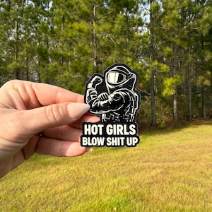 May include: A black and white sticker featuring a figure in a protective suit, flexing a bicep. The sticker has the text "HOT GIRLS BLOW SHIT UP" in white block letters on a black background.