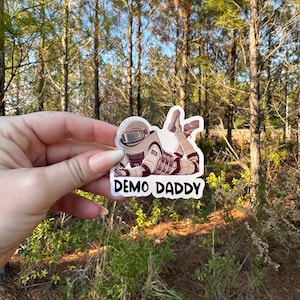 May include: A white sticker with a cartoon illustration of an astronaut in a white spacesuit lying on their stomach. The astronaut is wearing a helmet and has a brown belt. The text "DEMO DADDY" is printed in black below the astronaut.
