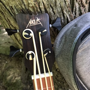 Op de afbeelding: Close-up van de kop van een zwarte en bruine akoestische basgitaar met de merknaam "AKLOT" gedrukt op de kop. De gitaar heeft vier snaren en zwarte stemknoppen.