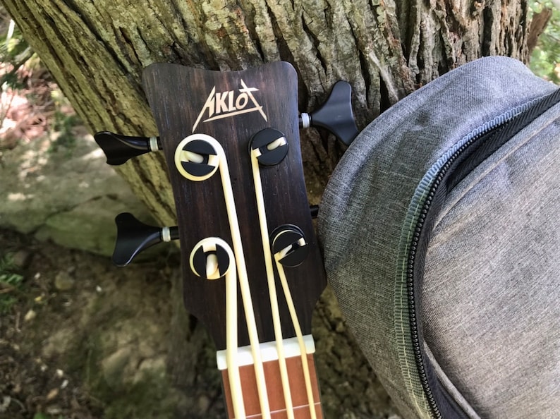 Op de afbeelding: Close-up van de kop van een zwarte en bruine akoestische basgitaar met de merknaam "AKLOT" gedrukt op de kop. De gitaar heeft vier snaren en zwarte stemknoppen.