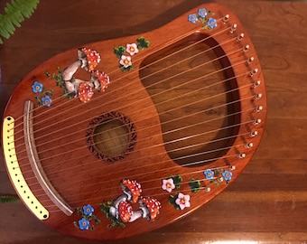 May include: A wooden lyre with a hand-painted design of mushrooms and flowers. The lyre has a gold-colored bridge and a sound hole with a decorative design.