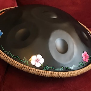 Op de afbeelding: Een zwarte handpan met een bruine touwrand en geschilderde bloemen in tinten blauw, roze en wit.
