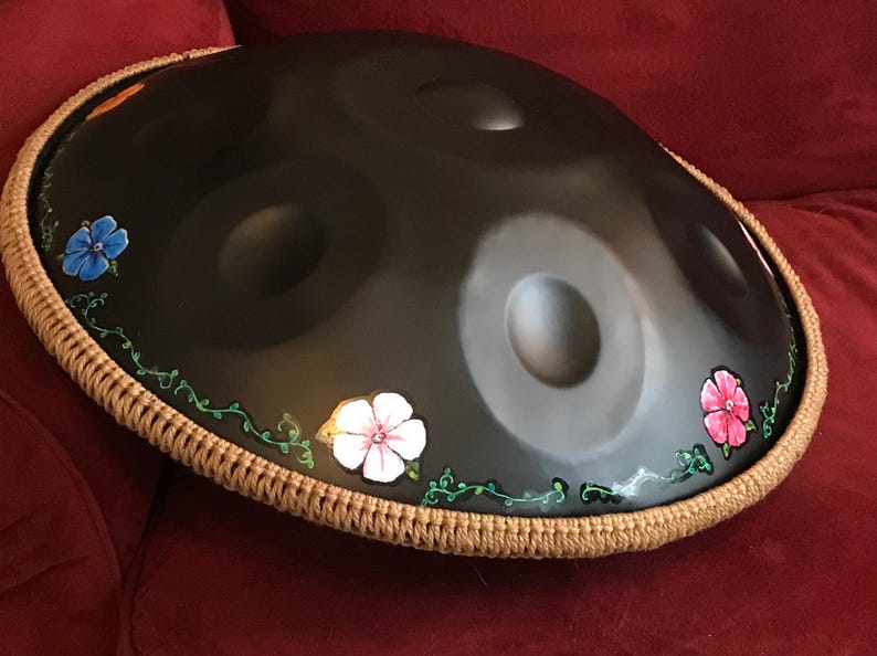 Op de afbeelding: Een zwarte handpan met een bruine touwrand en geschilderde bloemen in tinten blauw, roze en wit.