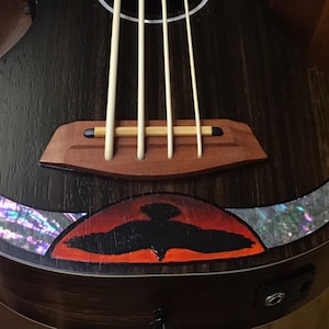 Op de afbeelding: Een donkerhouten ukulele met een zwart vogelontwerp en abalone-inlays. De ukulele heeft een houten brug en vier snaren.