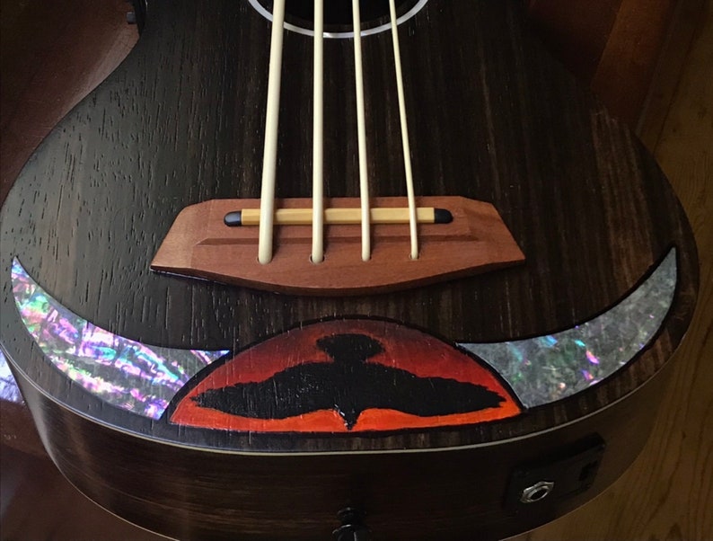 Op de afbeelding: Een donkerhouten ukulele met een zwart vogelontwerp en abalone-inlays. De ukulele heeft een houten brug en vier snaren.