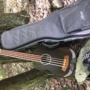 K&ouml;nnte beinhalten: Ein dunkelbrauner Ukulele-Bass mit einer floralen Einlage auf der Decke. Er liegt auf dem Boden neben einer grauen Ukulele-Tasche mit dem aufgestickten Wort "Acelot". Ein schwarz-wei&szlig; kariertes Band ist ebenfalls zu sehen.