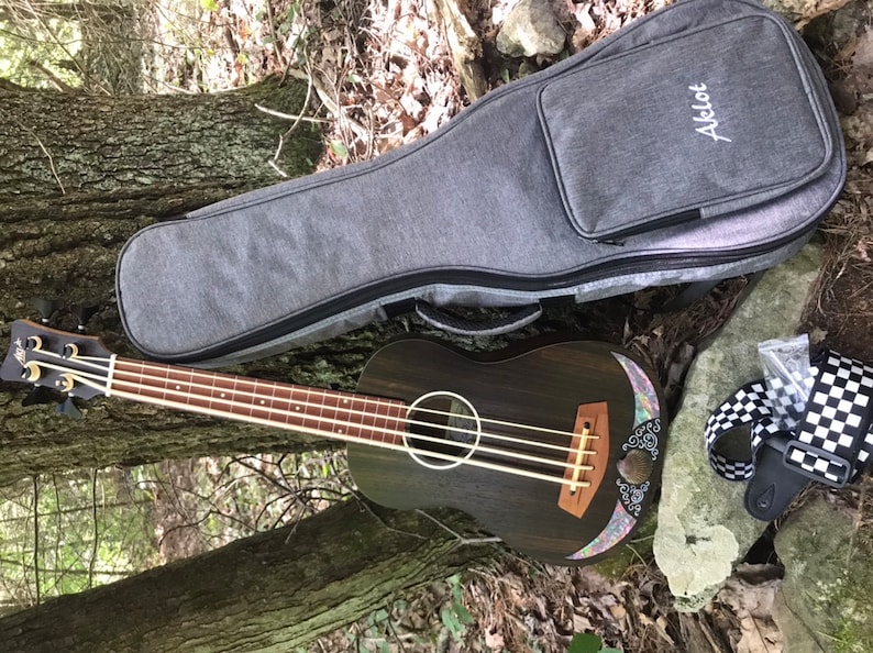 K&ouml;nnte beinhalten: Ein dunkelbrauner Ukulele-Bass mit einer floralen Einlage auf der Decke. Er liegt auf dem Boden neben einer grauen Ukulele-Tasche mit dem aufgestickten Wort "Acelot". Ein schwarz-wei&szlig; kariertes Band ist ebenfalls zu sehen.