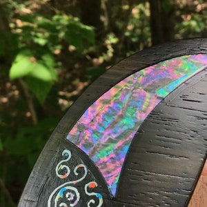 K&ouml;nnte beinhalten: Nahaufnahme einer dunklen Holzoberfl&auml;che mit einer dekorativen Einlage aus irisierendem Abalone-Perlmutt. Die Muschel ist sichelmondf&ouml;rmig und hat ein wirbelndes Muster in Rosa, Gr&uuml;n und Blau. Die Holzoberfl&auml;che ist auch mit wei&szlig;en Wirbeln verziert.