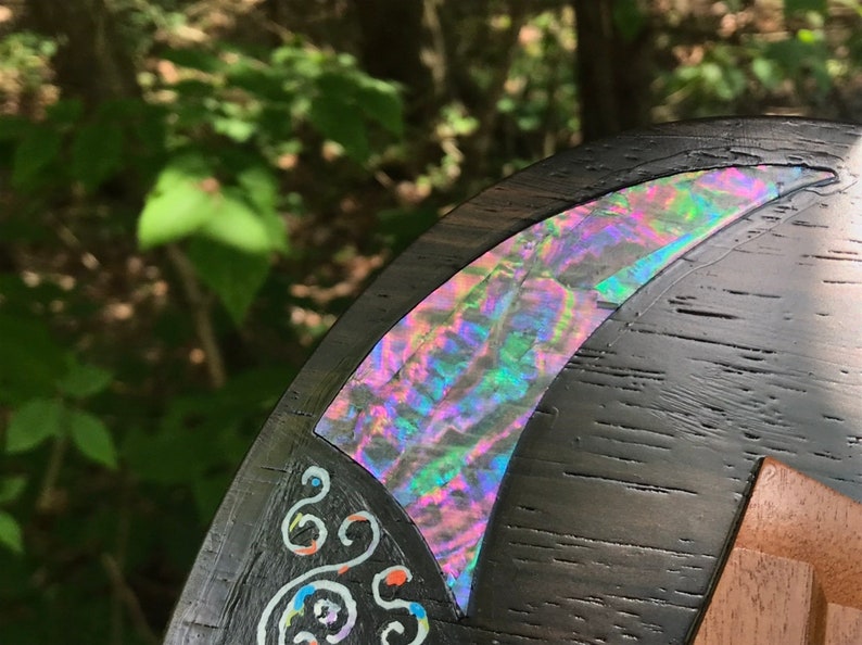 K&ouml;nnte beinhalten: Nahaufnahme einer dunklen Holzoberfl&auml;che mit einer dekorativen Einlage aus irisierendem Abalone-Perlmutt. Die Muschel ist sichelmondf&ouml;rmig und hat ein wirbelndes Muster in Rosa, Gr&uuml;n und Blau. Die Holzoberfl&auml;che ist auch mit wei&szlig;en Wirbeln verziert.