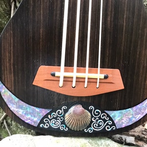 K&ouml;nnte beinhalten: Eine Ukulele aus dunklem Holz mit einer braunen Br&uuml;cke und einer Muscheleinlage. Die Einlage zeigt eine Muschel und ein wirbelndes Design.