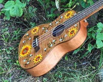 May include: A ukulele with a wood body and a sunflower design. The ukulele has a light brown body with a natural wood grain finish. The sunflower design is painted on the body of the ukulele and features yellow petals and brown centers. The ukulele is lying on a bed of green grass.