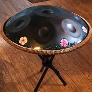 Op de afbeelding: Een zwarte handpan met een bruine geweven rand en roze bloemmotieven. De handpan staat op een zwarte metalen standaard.