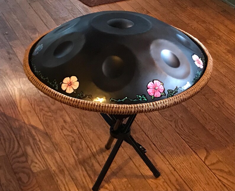 Op de afbeelding: Een zwarte handpan met een bruine geweven rand en roze bloemmotieven. De handpan staat op een zwarte metalen standaard.