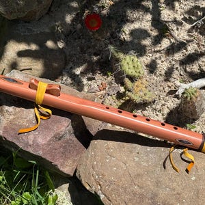 May include: A light brown wooden flute featuring a smooth cylindrical body, six finger holes, and a carved block secured with yellow leather ties. The instrument includes decorative inlays near both ends and additional yellow leather accents.