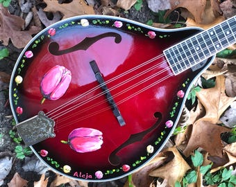 May include: A red, hand-painted mandolin with a black background and white and yellow floral accents. The mandolin has a floral design with pink tulips and the name "Aleja" written on the body.