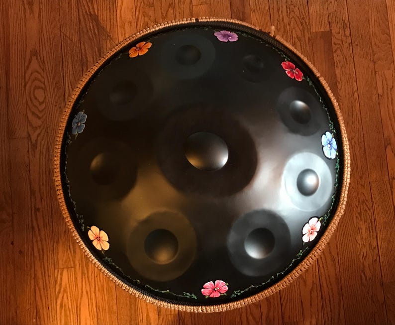 Op de afbeelding: Een zwarte stalen handpan met een bruine touwrand en geschilderde bloemen in verschillende kleuren rond de rand.