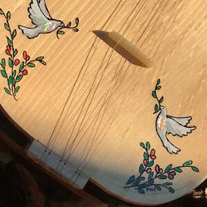 May include: A wooden musical instrument with a light wood finish. The instrument has a decorative design of two white doves with olive branches and red berries. The instrument is signed "Kori" on the bottom.