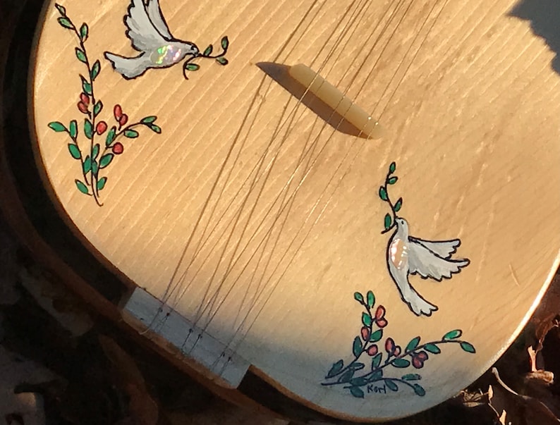 May include: A wooden musical instrument with a light wood finish. The instrument has a decorative design of two white doves with olive branches and red berries. The instrument is signed "Kori" on the bottom.