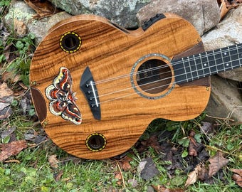 May include: A wooden acoustic ukulele with a natural grain finish, featuring a painted moth illustration on the body, two circular sound ports with yellow borders, a black bridge, a dark fretboard, and a central circular sound hole with inlay.