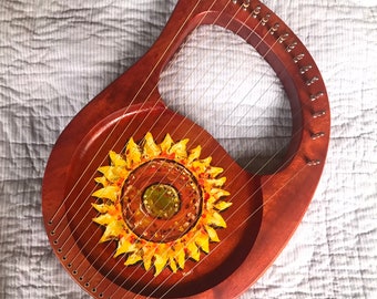 Può includere: Una lira in legno con un disegno di girasole dipinto sulla tavola armonica. La lira ha una finitura in legno naturale e piroli dorati.