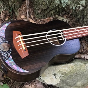K&ouml;nnte beinhalten: Ein dunkler Holzukelele-Bass mit einer Perlmutt-Einlage und einem Muschel-Design auf der Klangplatte. Die Ukulele liegt auf einem Stein und einigen Bl&auml;ttern.