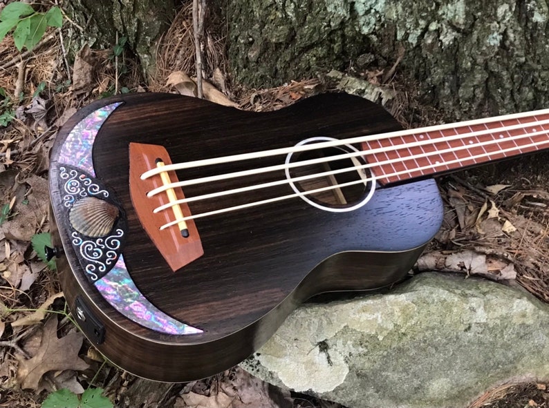 K&ouml;nnte beinhalten: Ein dunkler Holzukelele-Bass mit einer Perlmutt-Einlage und einem Muschel-Design auf der Klangplatte. Die Ukulele liegt auf einem Stein und einigen Bl&auml;ttern.