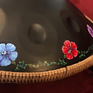 Op de afbeelding: Een zwarte handpan met een bruine gehaakte rand. De handpan is versierd met geschilderde bloemen in tinten blauw, rood en paars. De handpan is een muziekinstrument dat wordt bespeeld door met de vingers of handen op het oppervlak te slaan.