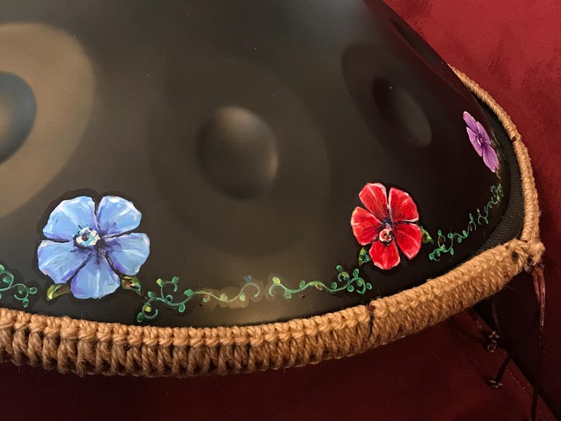 Op de afbeelding: Een zwarte handpan met een bruine gehaakte rand. De handpan is versierd met geschilderde bloemen in tinten blauw, rood en paars. De handpan is een muziekinstrument dat wordt bespeeld door met de vingers of handen op het oppervlak te slaan.