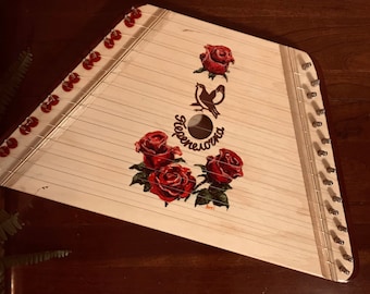 Può includere: Un'arpa a corde in legno con rose dipinte in rosso e bianco e un disegno di un uccello. L'arpa ha un foro sonoro e un'etichetta che recita "Taeponeiorna".
