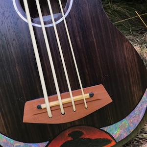 Op de afbeelding: Close-up van een donkerhouten ukulele met een zwart vogelontwerp op de onderste bout. De ukulele heeft een houten brug en een witte abalone-inleg rond de klankgat.