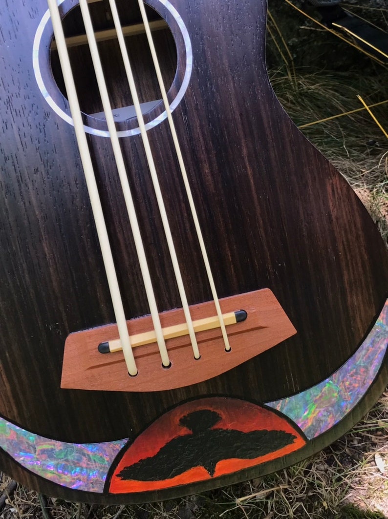 Op de afbeelding: Close-up van een donkerhouten ukulele met een zwart vogelontwerp op de onderste bout. De ukulele heeft een houten brug en een witte abalone-inleg rond de klankgat.