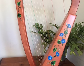 May include: A wooden harp with a natural finish and blue flower designs painted on the body. The harp has strings and tuning pegs.