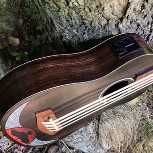 Op de afbeelding: Een donkerbruine houten ukulele met een witte pickguard en een zwarte pickup. De ukulele heeft een halve maan inleg op het klankgat en een rood en zwart vogelontwerp.