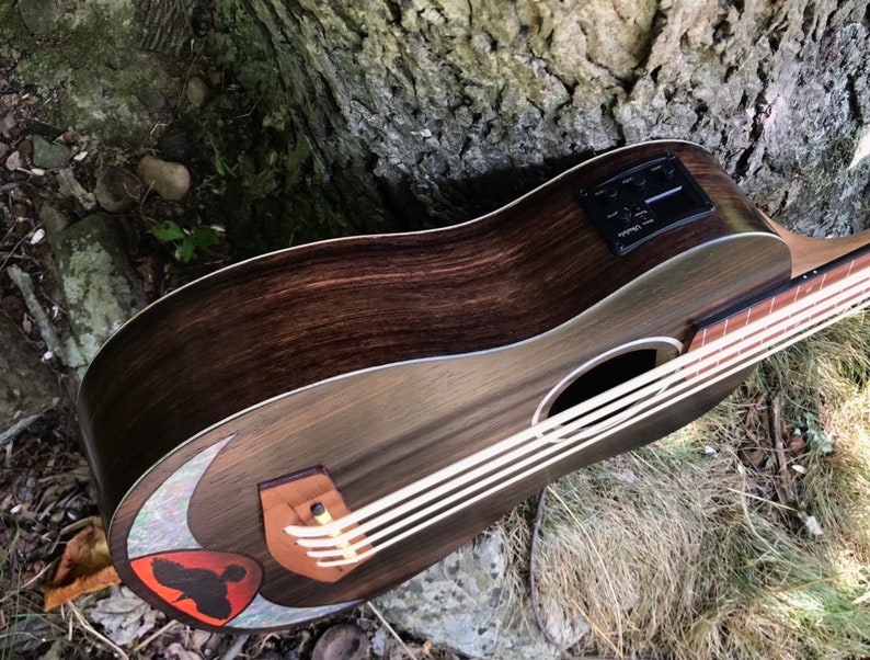 Op de afbeelding: Een donkerbruine houten ukulele met een witte pickguard en een zwarte pickup. De ukulele heeft een halve maan inleg op het klankgat en een rood en zwart vogelontwerp.