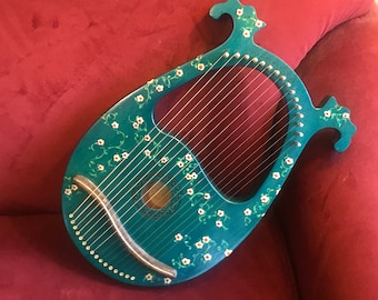 May include: A teal blue lyre with white flower designs painted on the body. The lyre has a curved soundboard and a set of strings.