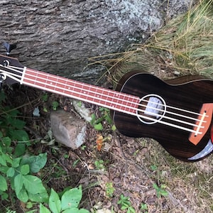Op de afbeelding: Een zwarte ukulelebas met een houtnerf afwerking en een rood, wit en blauw inleg in het klankgat. De bas ligt op een bed van groen gras en bruine bladeren.