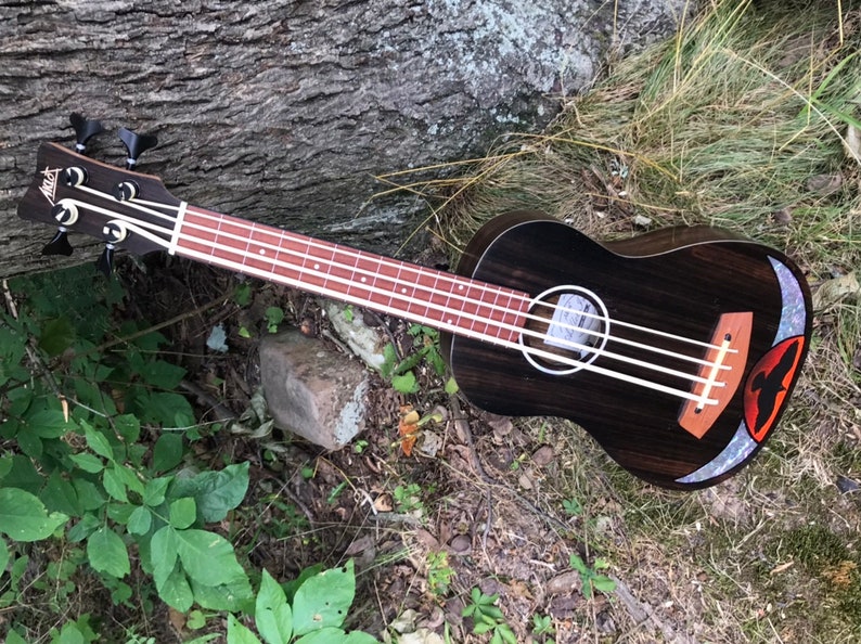 Op de afbeelding: Een zwarte ukulelebas met een houtnerf afwerking en een rood, wit en blauw inleg in het klankgat. De bas ligt op een bed van groen gras en bruine bladeren.