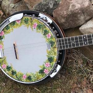May include: A five-string banjo with a painted design of green shamrocks and pink flowers around the rim. The banjo has a chrome-plated resonator and a wooden neck.