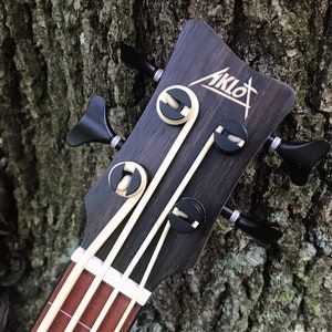 K&ouml;nnte beinhalten: Nahaufnahme des Kopfes eines schwarzen Ukulelen-Basses mit einer wei&szlig;en Einlage. Der Kopf hat vier Stimmwirbel mit schwarzen Kn&ouml;pfen und ein wei&szlig;es Logo mit der Aufschrift "Aklot".