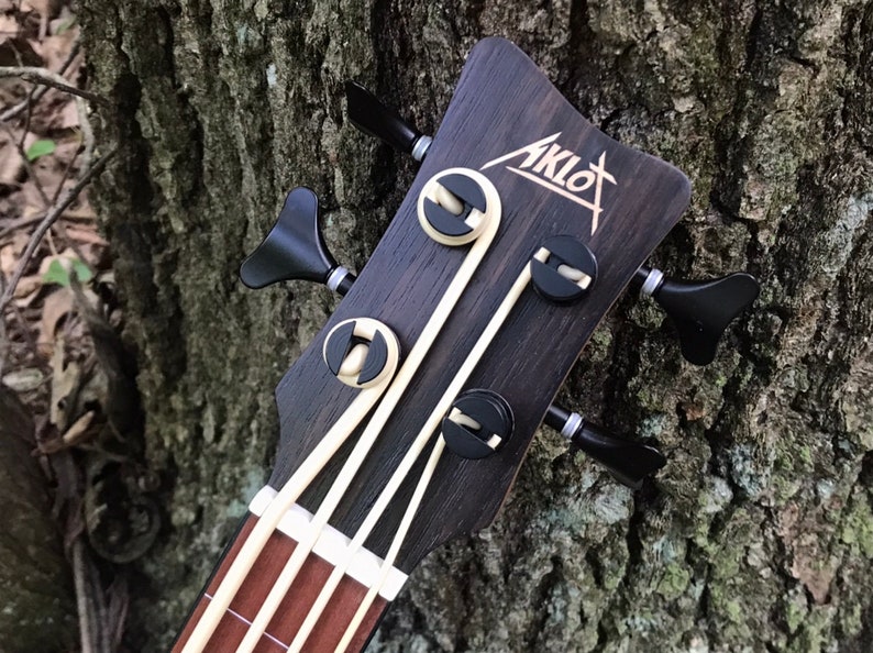 K&ouml;nnte beinhalten: Nahaufnahme des Kopfes eines schwarzen Ukulelen-Basses mit einer wei&szlig;en Einlage. Der Kopf hat vier Stimmwirbel mit schwarzen Kn&ouml;pfen und ein wei&szlig;es Logo mit der Aufschrift "Aklot".
