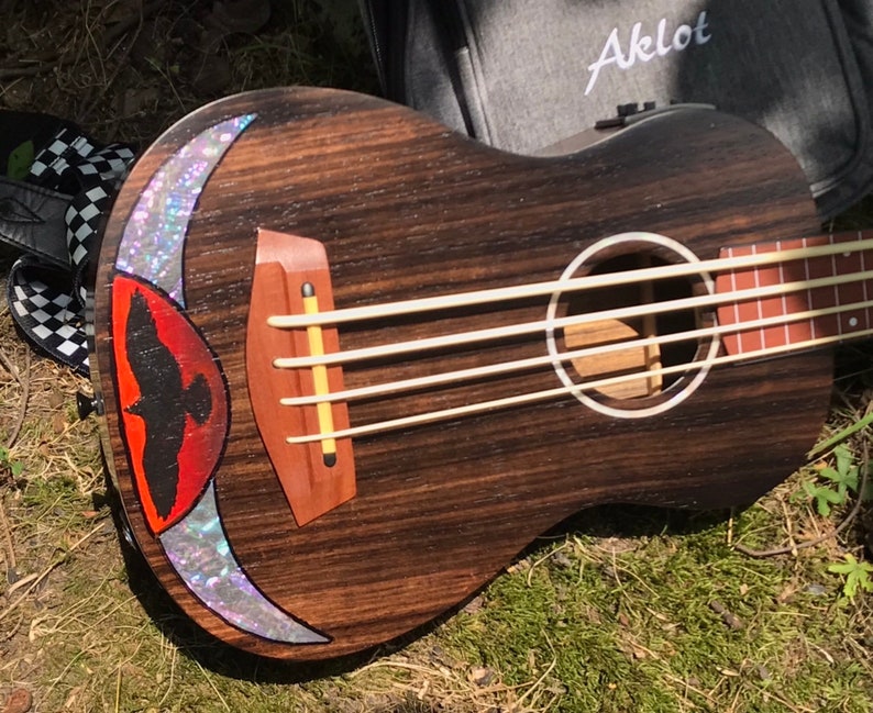 Op de afbeelding: Een donkerbruine ukulelebas met een rood en wit vogelontwerp op het lichaam. De bas heeft vier snaren en een klankgat met een witte ring eromheen. De bas is gemaakt door Aklot.